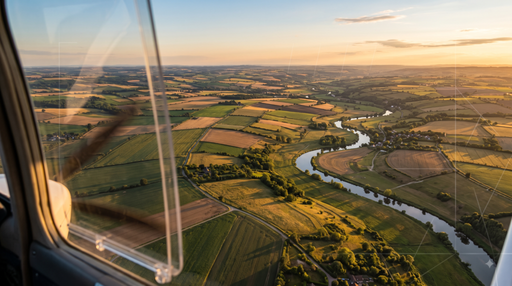 Photorealistic aerial photograph of farmland and a river seen from a survey aircraft during a mapping flight, with faint flight-line grid lines overlaid.